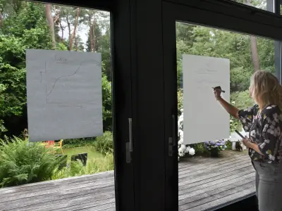 Buiten vergaderen op het terras: iemand schrijft op een flip-over, gezien door de openslaande deuren van de vergaderruimte