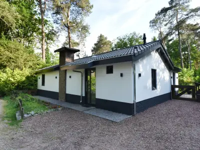 Witte bungalow met zwarte plint en schoorsteen, omgeven door bos en een grindpad