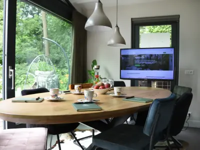 Ovale houten vergadertafel gedekt met koffie, fruitwater en notitieboekjes, met schuifpui naar de bostuin en een groot scherm aan de wand